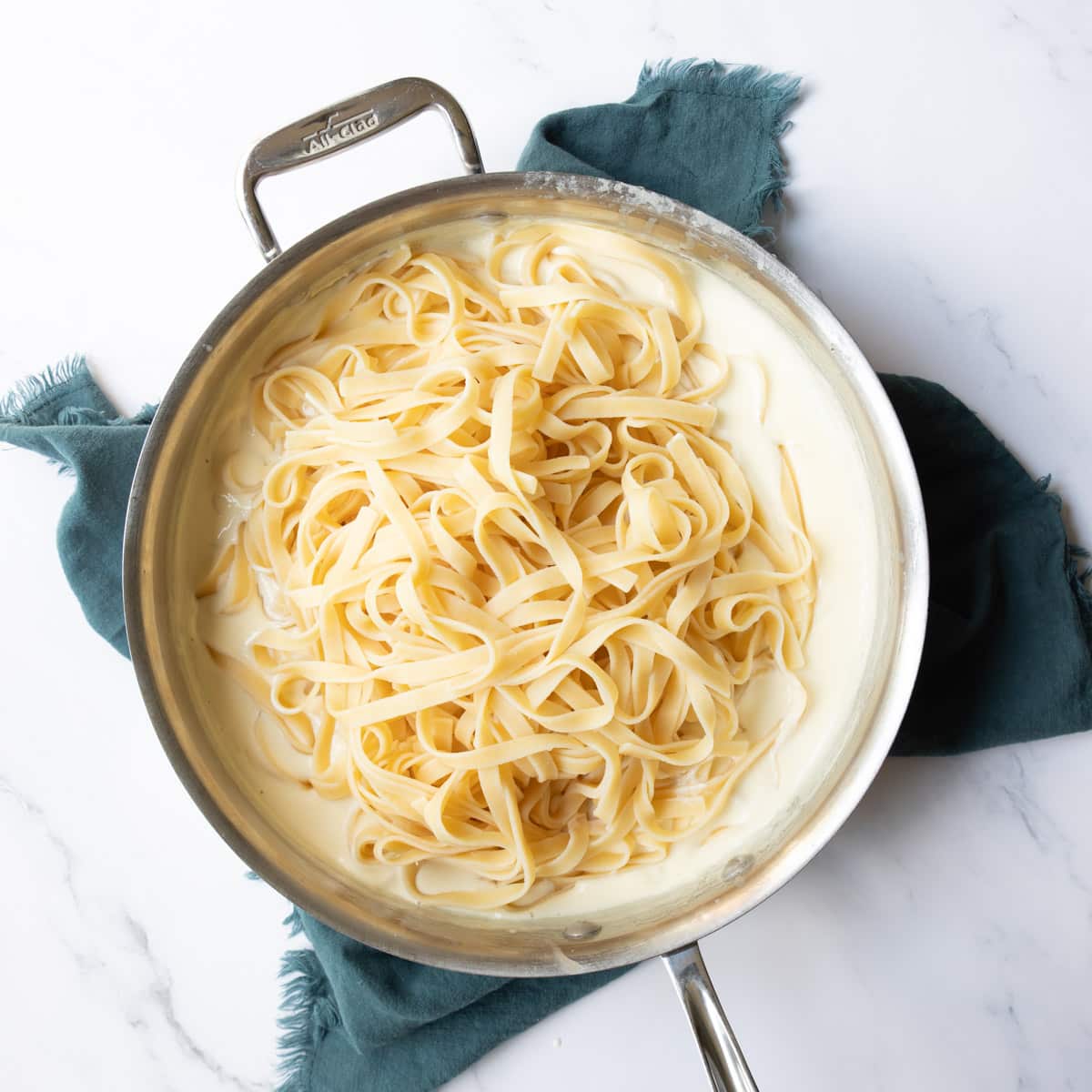 Freshly cooked linguine noodles added to the top of a saucepan filled with rich creamy white alfredo sauce.