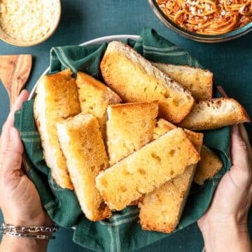 Hands picking up a bowl of baked garlic bread cut into thin slices laying on a decorative towel.