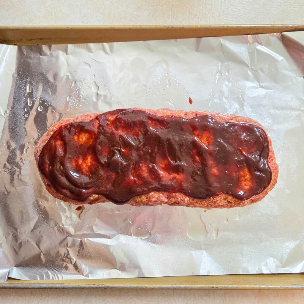Barbecue glaze covering a raw beef meatloaf loaf ready to bake.