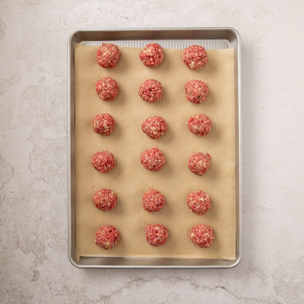 Uniform sized raw meatballs laying on parchment paper on a baking sheet.