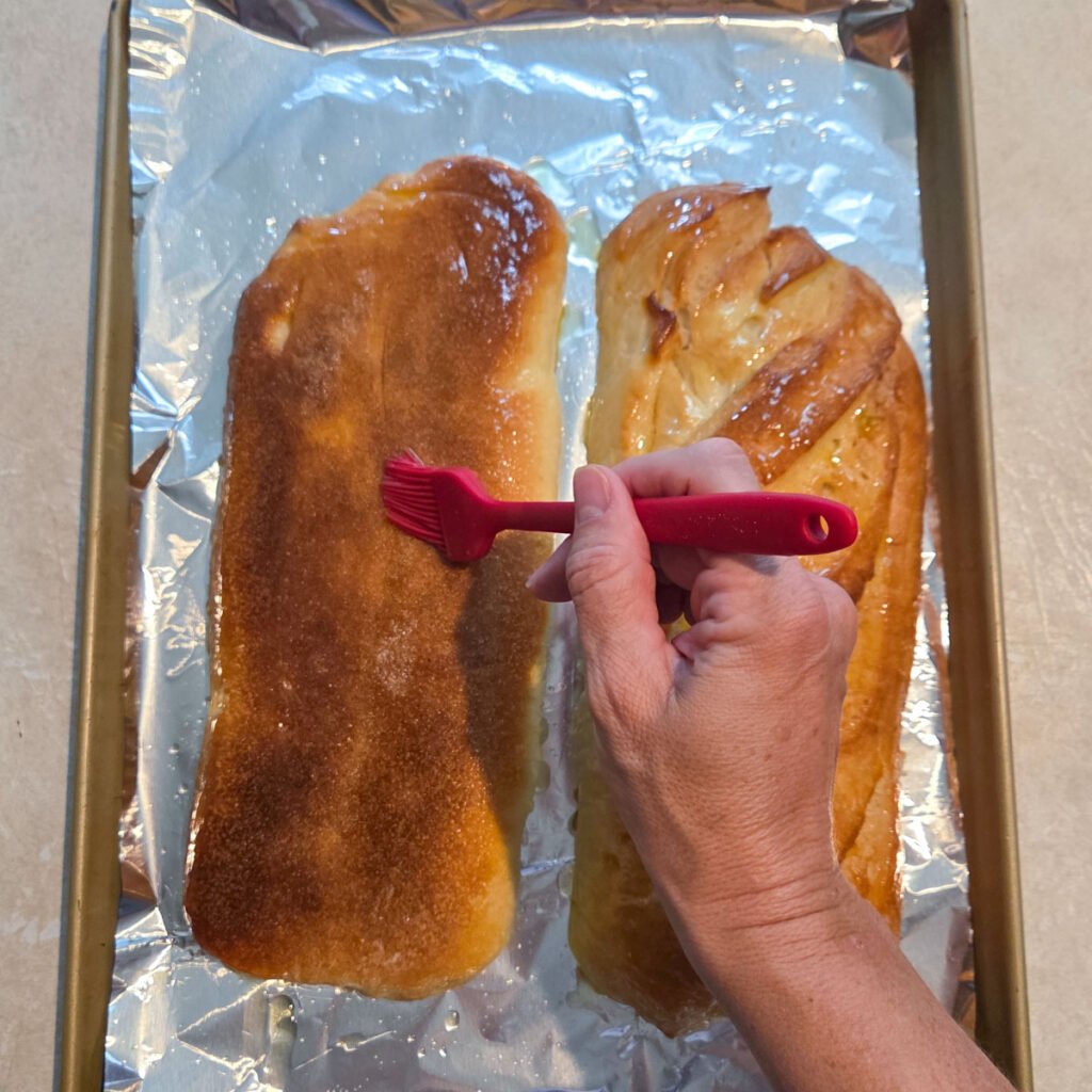 Butter being brushed over the top of halved French bread loaves on a foil lined baking sheet.