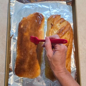 Butter being brushed over the top of halved French bread loaves on a foil lined baking sheet.