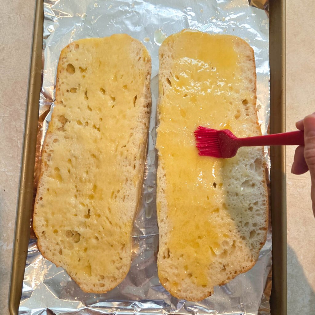 Melted butter being brushed over French bread loaf with a silicone brush that's cut in half.