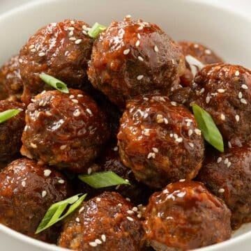 Bowl of sweet and sour meatballs garnished with sesame seeds and sliced green onions.