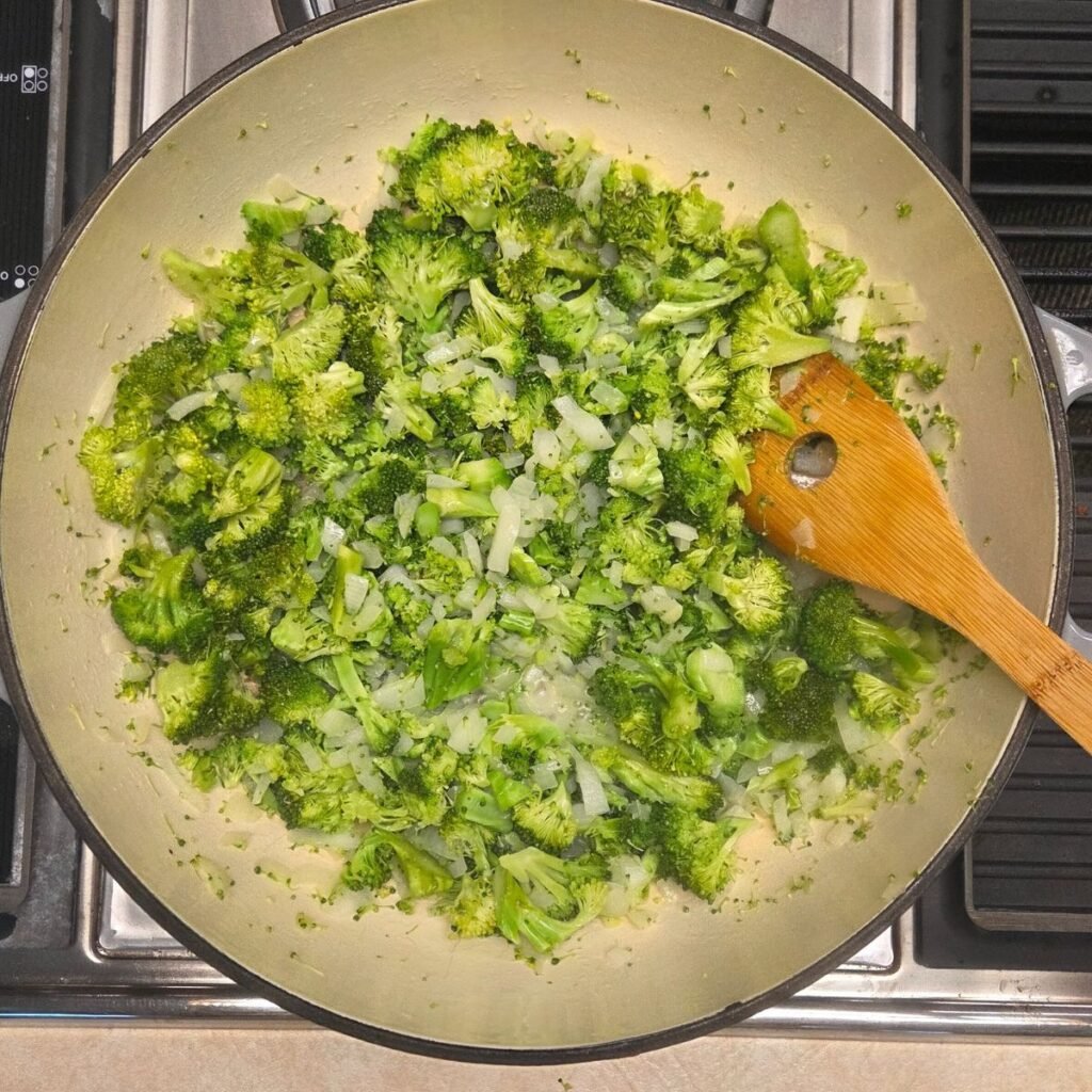 Wooden spoon sauteeing onions and broccoli florets in a Dutch oven pan.