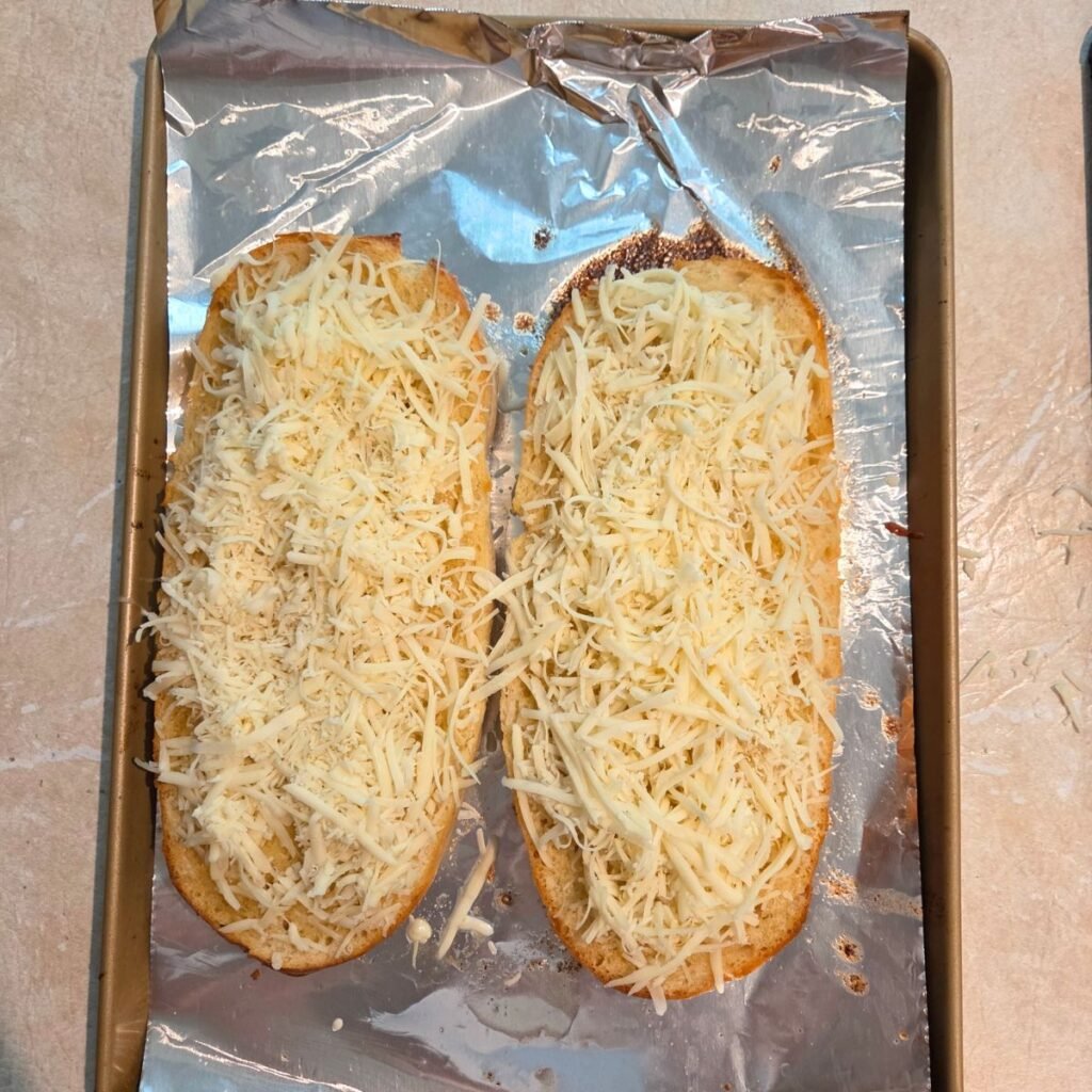Shredded cheese added to the two halves of a garlic butter topped loaf of Italian bread.