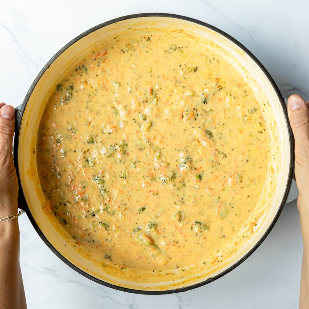 Hands picking up a Dutch oven pan filled with creamy Broccoli cheddar soup.