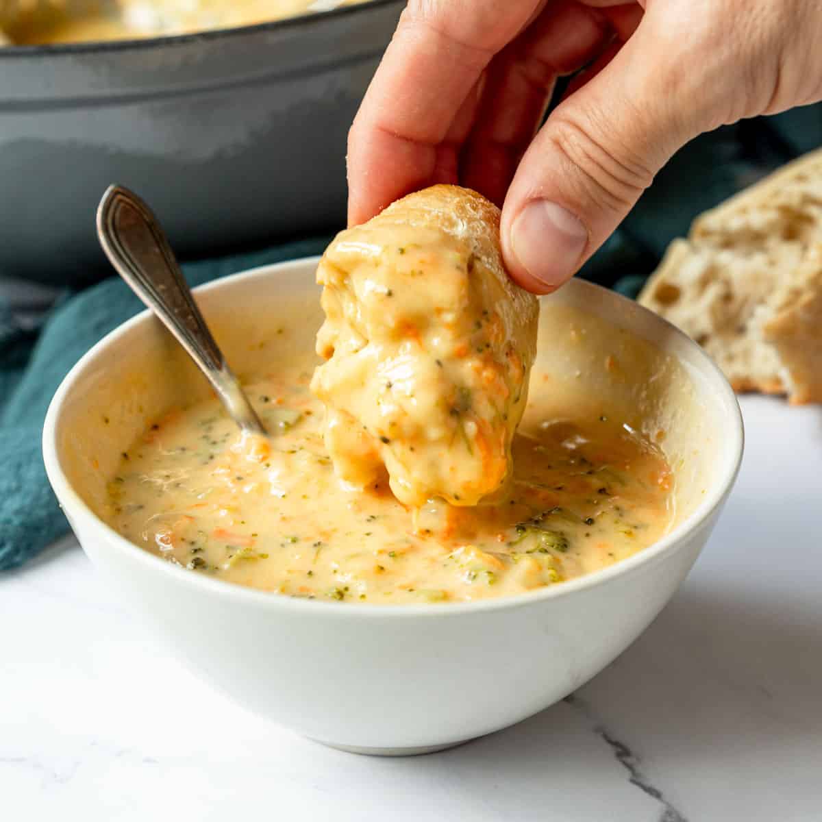 Hand dunking a hunk of bread into a bowl of with creamy broccoli cheddar soup.