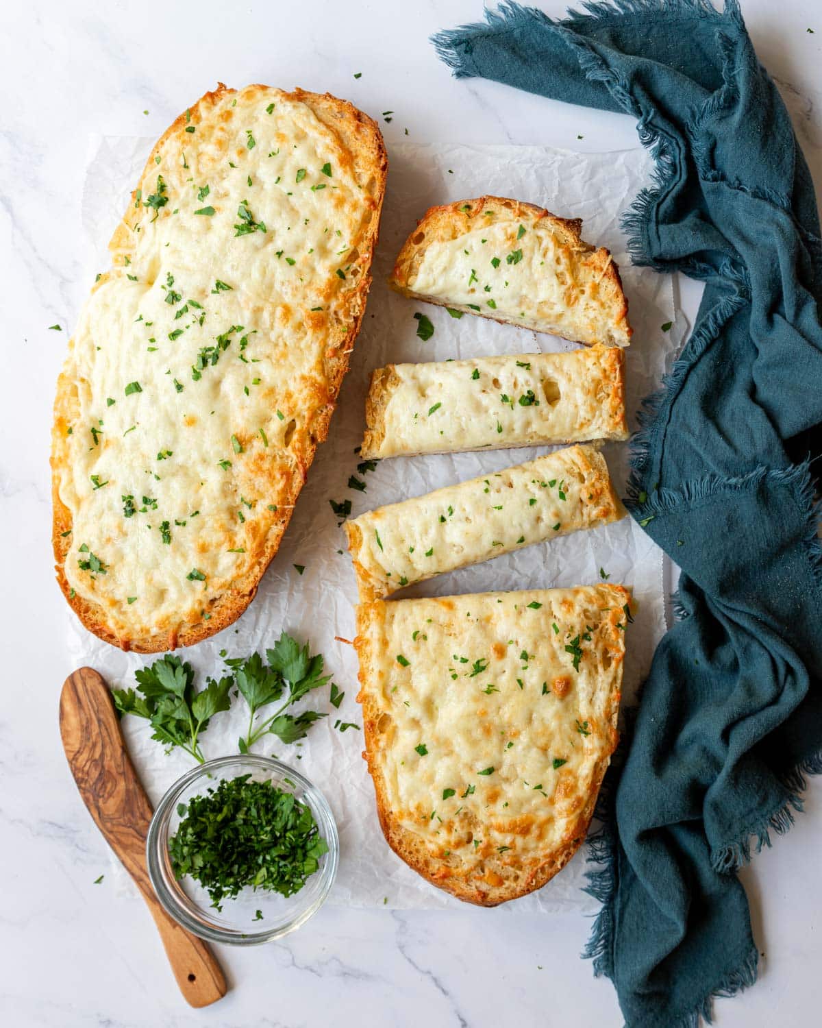 Cheesy garlic bread with half a loaf sliced up ready to serve next to another golden brown cheesy bread loaf garnished with chopped up parsley.