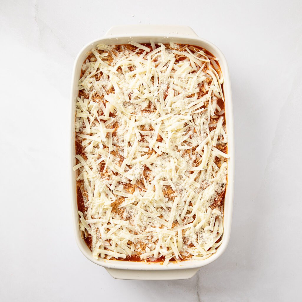 Shredded cheese topping the cheese stuffed manicotti shells in a baking dish.