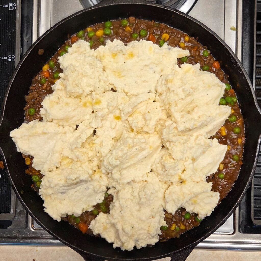 Dollops of mashed potatoes added to a skillet with beef and veggie mixture.