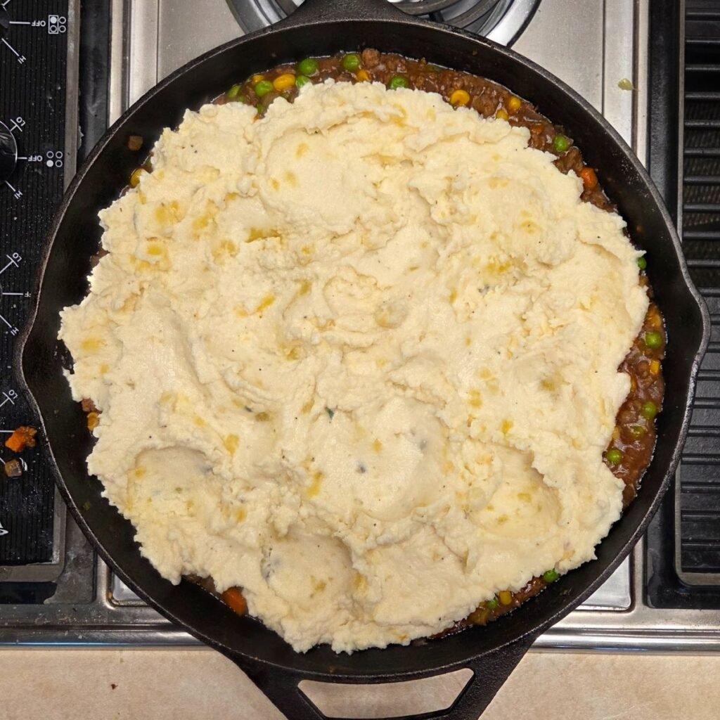 Buttery mashed potatoes added to a skillet with ground beef gravy and veggies mixture, ready to bake.