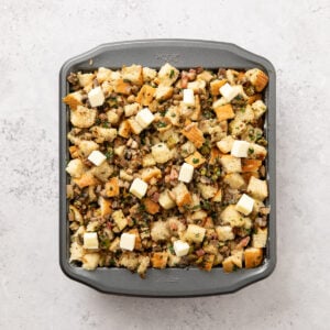 Square 8x8 baking pan with mushroom stuffing in a pan and topped with cubes of butter, ready to bake.