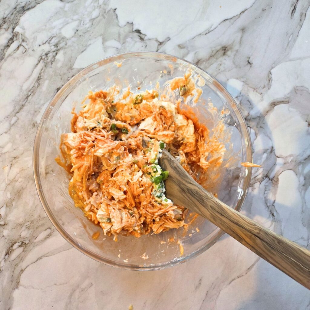 Chunk chicken and the ingredients for making buffalo dip being combined in a glass mixing bowl.