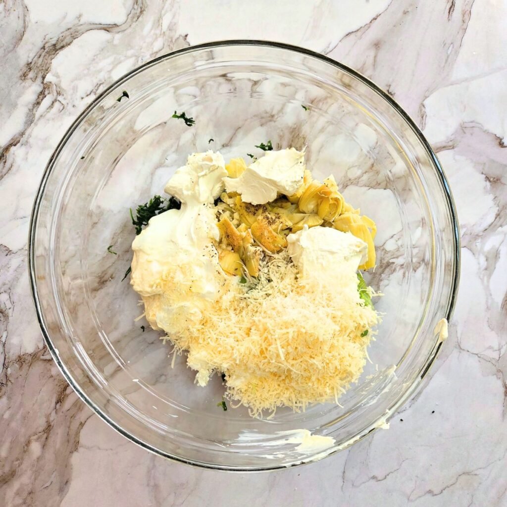 Artichokes and ingredients added together in a glass bowl before combining.
