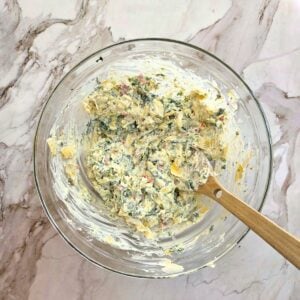 Wooden spoon resting in a bowl of creamy spinach artichoke dip ingredients before baking.