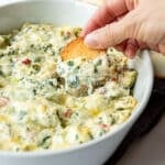 Hand dipping a bagel chip in for a scoop of baked spinach artichoke dip.