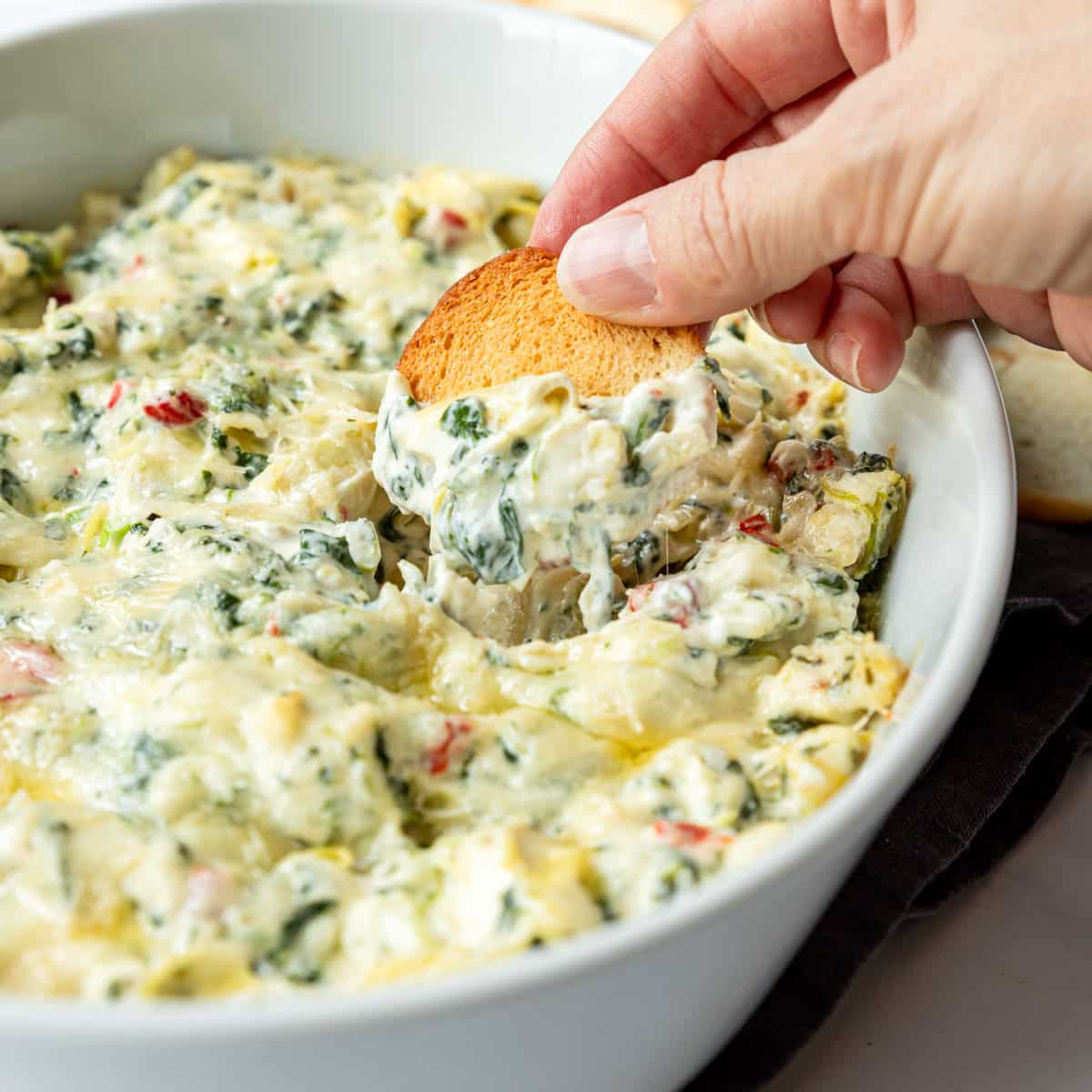 Hand dipping a bagel chip in for a scoop of baked spinach artichoke dip.