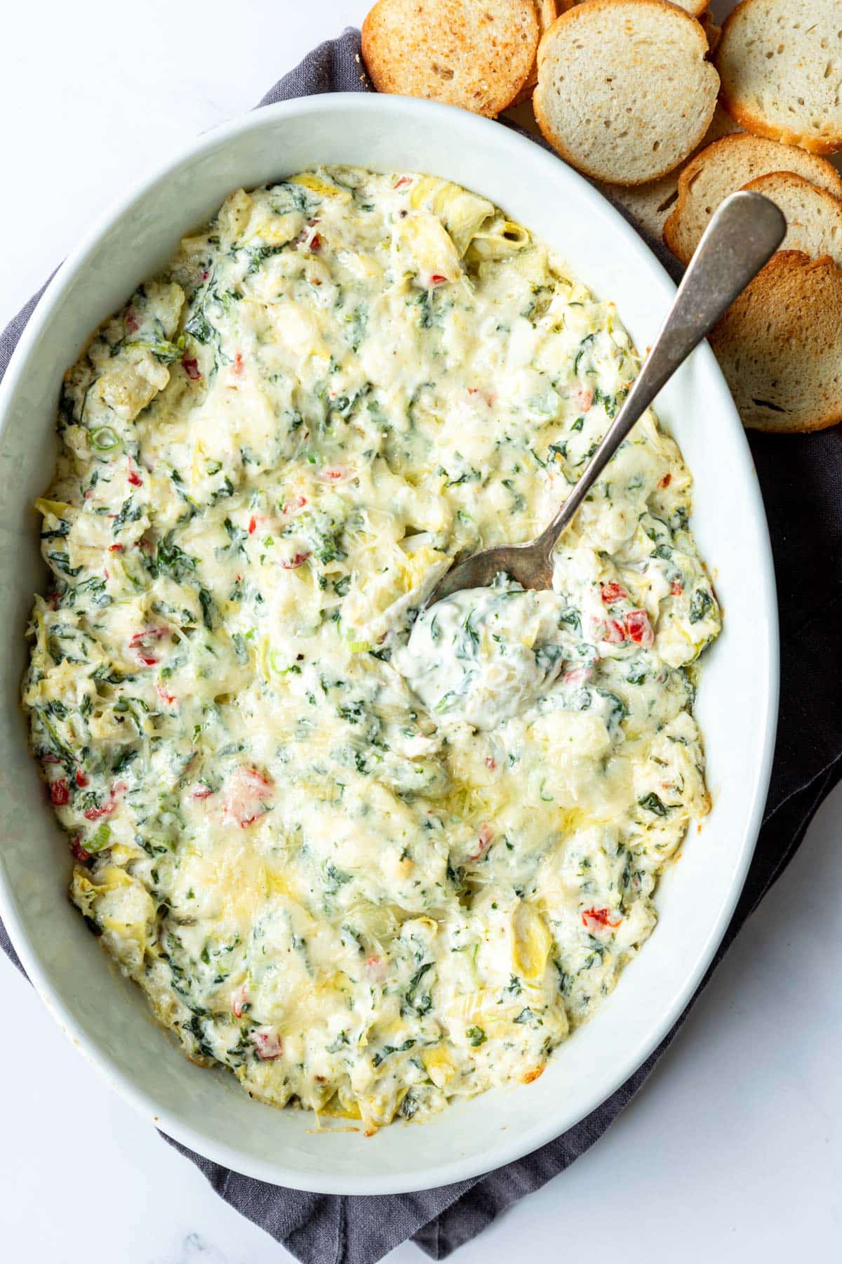 A spoon resting in an oval baking dish of creamy spinach artichoke dip.