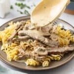 Wooden spoon dripping stroganoff sauce onto a plate of pasta and steak.