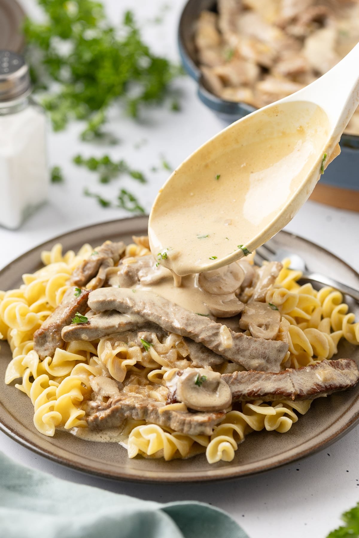 Beef stroganoff served with mushroom pieces over a bed of pasta with creamy sauce being spooned on the plate.
