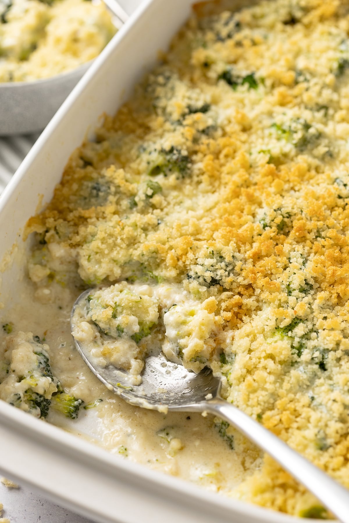 Spoon resting in a serving dish for broccoli cheese casserole dish with a portion gone.