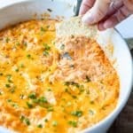 Hand dipping a tortilla chip into a golden baked buffalo chicken dip topped with thin chopped chives.