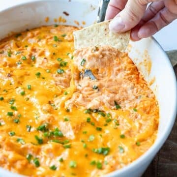 Hand dipping a tortilla chip into a golden baked buffalo chicken dip topped with thin chopped chives.