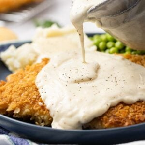 Close up of homemade gravy being poured over a crispy crunchy fried steak ready to eat with potatoes and peas.