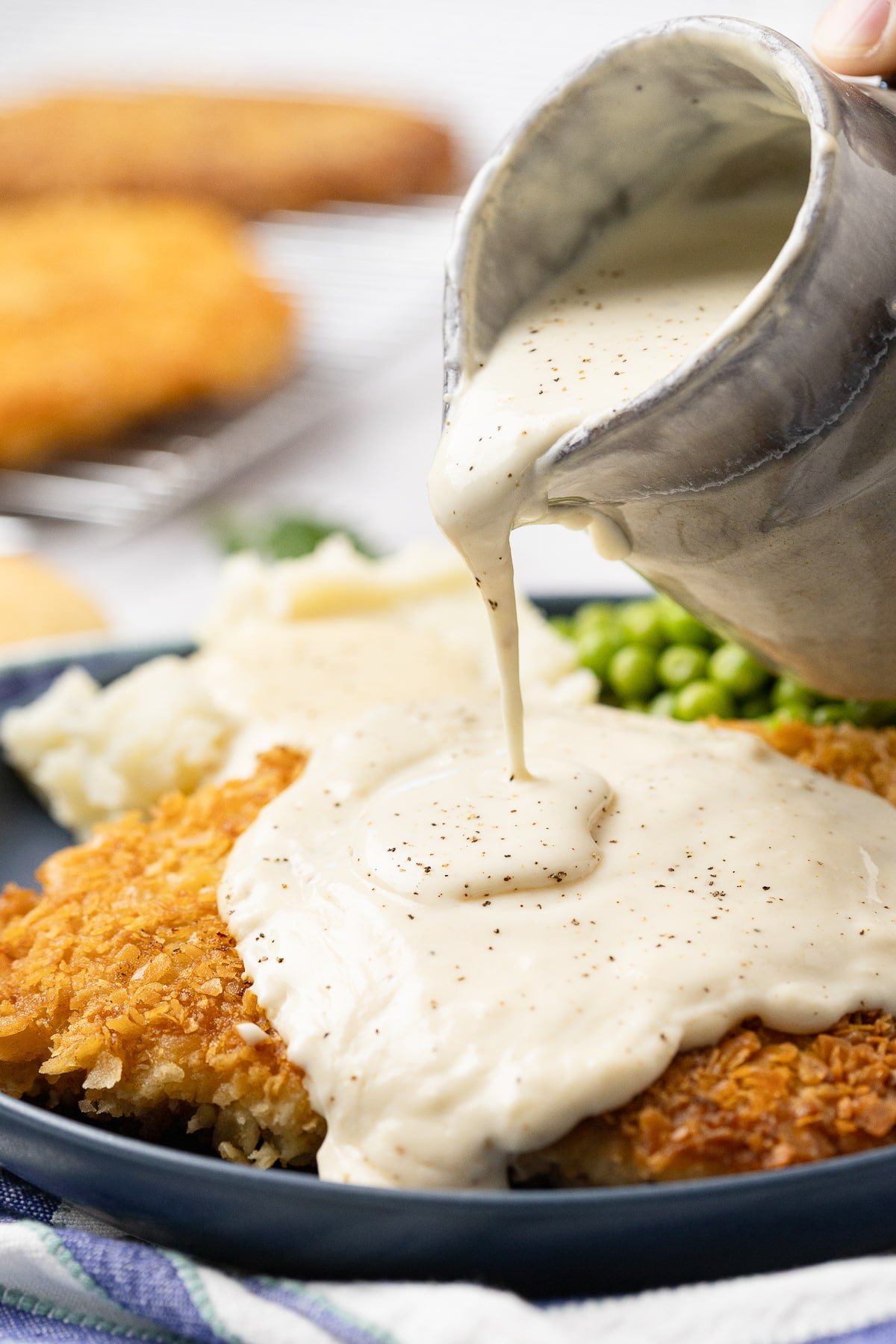 Homemade gravy being poured over a crispy country fried steak on a plate served with mashed potatoes and peas.