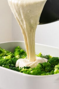 Homemade white cheese sauce being poured over bright green broccoli florets in a baking dish.