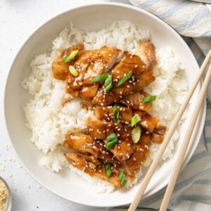 Chopsticks resting on the edge of a bowl served with rice, and chicken thighs topped with teriyaki sauce.