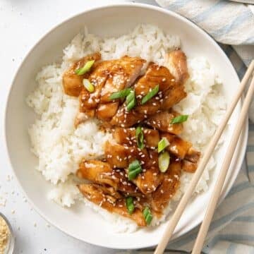 Chopsticks resting on the edge of a bowl served with rice, and chicken thighs topped with teriyaki sauce.