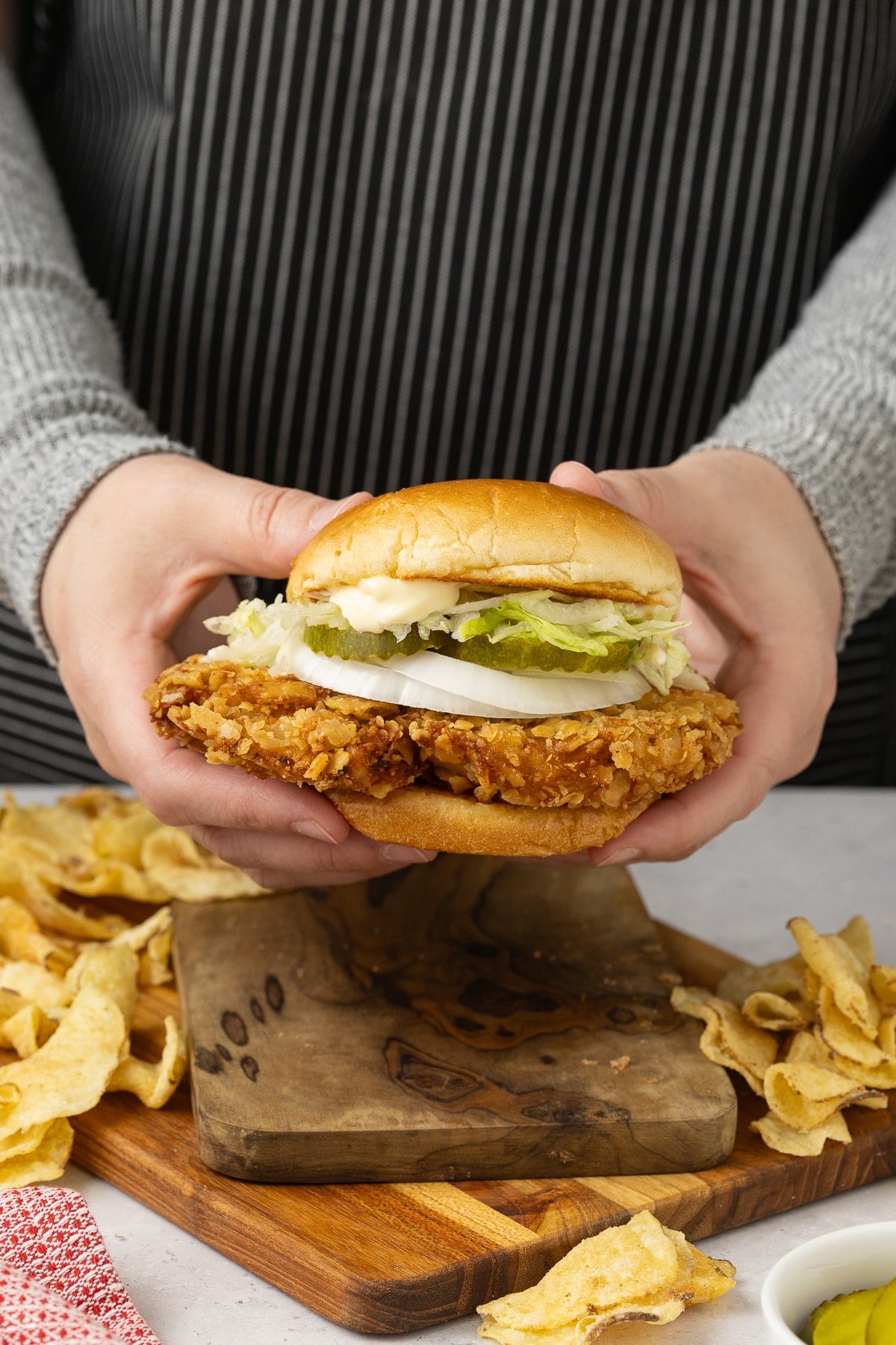 Two hands holding a fried pork tenderloin sandwich with chips laying around under the sandwich.