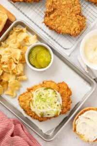 Top view of a baking tray that's served a fried pork tenderloin sandwich topped with horseradish mayo, shredded lettuce, and pickles with chips on the side.
