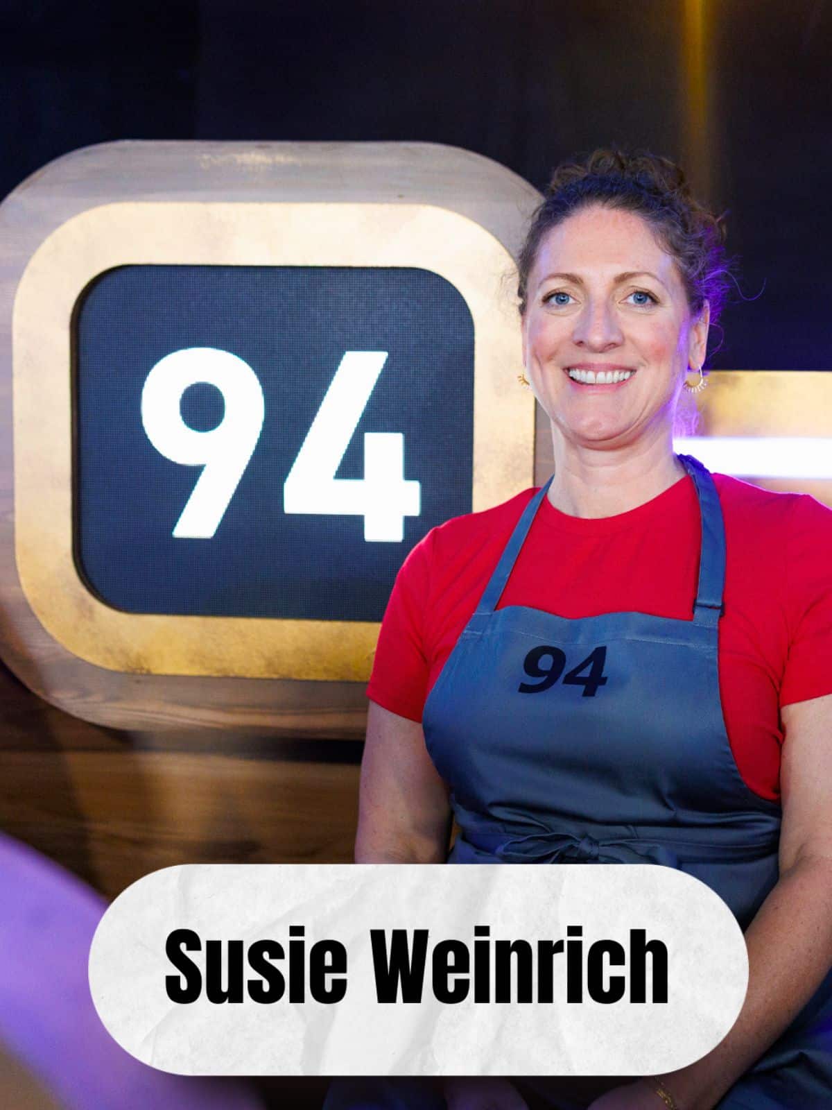 Susie Weinrich in a red shirt and apron with the number 94 on the front, sitting on a Food Network set, in front of the #94