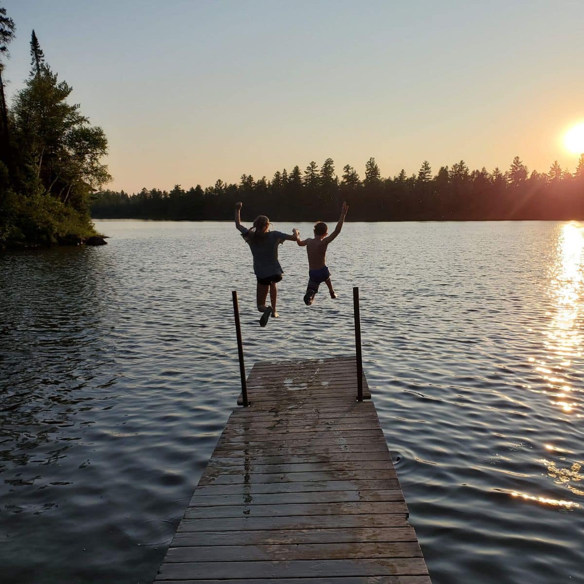 two kids jumping in a lake off the dock at sunset 