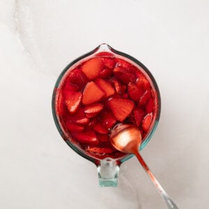 Spoon coming out of jello in a glass measuring bowl with sliced strawberries floating in it.