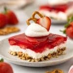 Dollop of whipped cream garnished with a slice of fresh strawberry and pretzel on top of a square serving of strawberry pretzel salad.