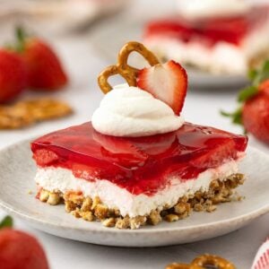 Dollop of whipped cream garnished with a slice of fresh strawberry and pretzel on top of a square serving of strawberry pretzel salad.