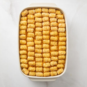 Frozen tater tots arranged over cheese, gravy, ground beef and mixed veggies in a baking dish, ready to cook.